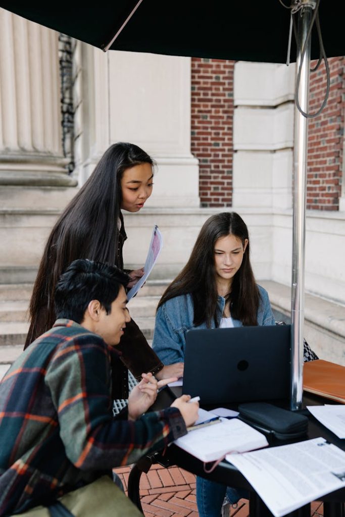 A group of university students collaborating on a project outdoors under an umbrella, laptops and notebooks in use.
